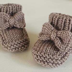 May include: A pair of knitted baby booties in a soft taupe color. Each bootie features a decorative bow on the front. The booties are made of a textured yarn, perfect for keeping little feet warm.