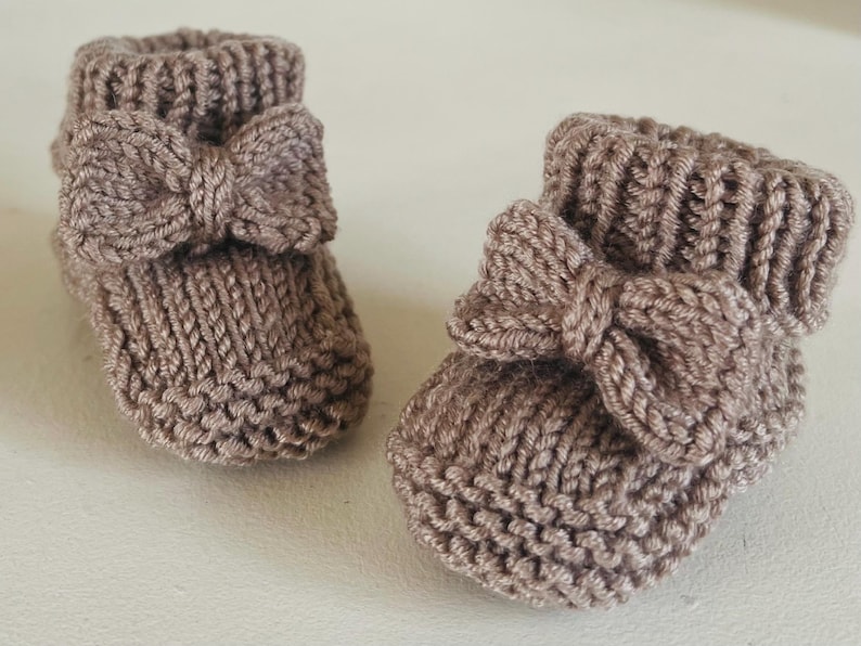 May include: A pair of knitted baby booties in a soft taupe color. Each bootie features a decorative bow on the front. The booties are made of a textured yarn, perfect for keeping little feet warm.