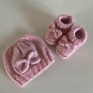May include: A matching set of baby accessories in a soft pink hue. The set includes a knitted beanie hat and a pair of booties, each adorned with a decorative bow. The items are crafted from a textured yarn, suggesting a handmade or artisanal quality.