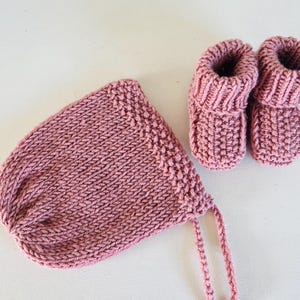 Pode incluir: Um conjunto de bebé tricotado em cor rosa empoeirado. O conjunto inclui um gorro com laços e um par de botinhas. Os artigos são feitos de um fio macio e texturizado, perfeito para um recém-nascido. O gorro tem cerca de 15 cm de largura.
