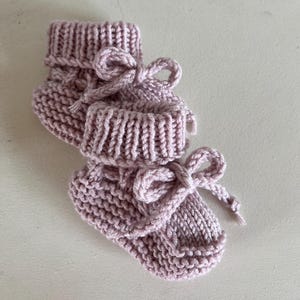 May include: A pair of hand-knitted baby booties in a soft, muted pink. The booties feature a ribbed cuff and are tied with a matching yarn bow. The knitted texture is visible, showcasing the craftsmanship of the item.