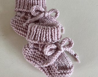 May include: A pair of dusty pink hand-knitted baby booties featuring a garter stitch texture, a ribbed cuff, and a decorative bow tied at the front. The booties are crafted from a soft yarn and display a consistent knit pattern throughout the design.