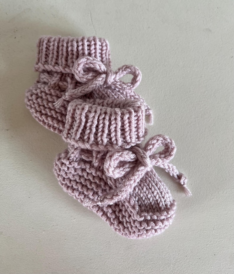 Puede incluir: Un par de patucos de beb&eacute; tejidos a mano en un rosa suave y apagado. Los patucos tienen un pu&ntilde;o acanalado y est&aacute;n atados con un lazo de hilo a juego. La textura tejida es visible, mostrando la artesan&iacute;a del art&iacute;culo.