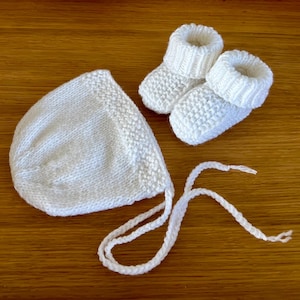 May include: A white knitted baby set, including a bonnet with ties and a pair of booties. The set is made of soft, textured yarn. The items are displayed on a wooden surface.