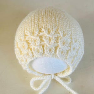 May include: A cream-coloured knitted baby bonnet featuring a floral lace design on the front. The bonnet has a rounded shape and a tie closure under the chin. The soft, textured knitwear is suitable for newborns.