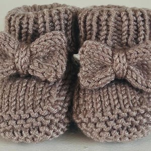May include: A pair of knitted baby booties in a soft taupe color. Each bootie features a decorative bow on the front. The booties are made with a thick knit stitch and have a ribbed cuff.