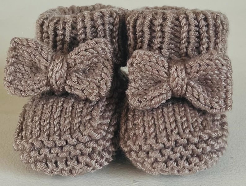 May include: A pair of knitted baby booties in a soft taupe color. Each bootie features a decorative bow on the front. The booties are made with a thick knit stitch and have a ribbed cuff.