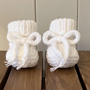 Op de afbeelding: Een paar witte gebreide babyslofjes. Elk slofje heeft een strik aan de voorkant. De slofjes zijn gemaakt van zacht, getextureerd garen en zijn ontworpen om de voetjes van een baby warm te houden. Gepresenteerd op een houten oppervlak.