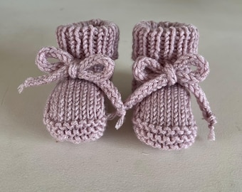 May include: A pair of dusty rose knitted booties featuring a ribbed cuff, a garter stitch toe section, and a matching knitted tie threaded through the ankle area, tied into a bow at the front of each bootie, showcasing a textured yarn construction.