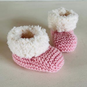 May include: A pair of baby booties. The booties are knit in a light pink yarn with a fluffy white trim around the top. The booties are designed to keep a baby's feet warm and cozy.