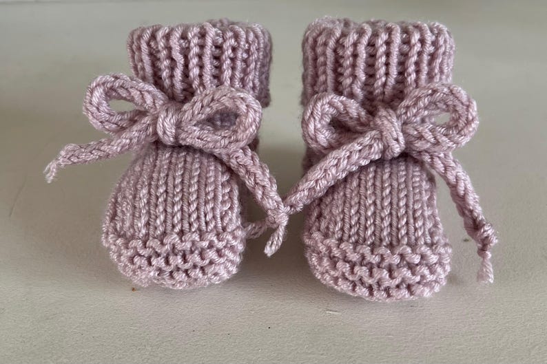 Puede incluir: Un par de patucos de beb&eacute; tejidos en color lavanda suave. Cada patuco presenta un lazo decorativo en el tobillo. Los patucos est&aacute;n hechos de un hilo texturizado, perfecto para un reci&eacute;n nacido.
