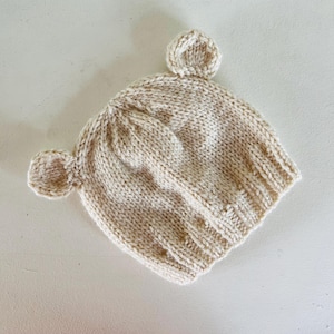 May include: A cream-colored knit beanie hat featuring two small, rounded ears attached to the top sides. The hat is constructed with a textured stockinette stitch pattern and includes a ribbed brim at the base, crafted from soft yarn.