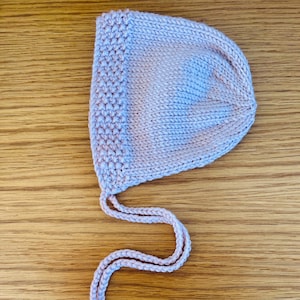 May include: A light pink knitted baby bonnet with a rounded shape and attached ties. The bonnet is made of soft yarn and features a textured border. The ties are designed to secure the bonnet under the chin.