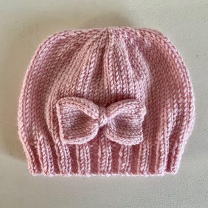 May include: A light pink knitted beanie hat featuring a ribbed texture and a matching knitted bow centred on the front. The hat is constructed with a thick yarn knit pattern, showcasing a rounded crown and a structured, folded brim edge.