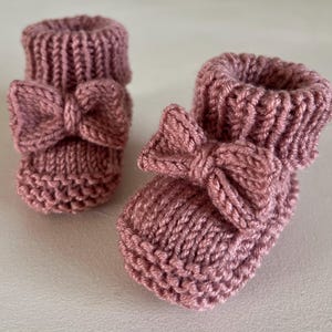 May include: A pair of knitted baby booties in a dusty rose color. Each bootie features a decorative bow on the front. The booties are made of a soft, textured yarn, perfect for a newborn.