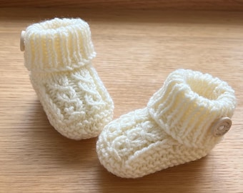 May include: A pair of knitted baby booties in a cream colour with a wooden button on each bootie.