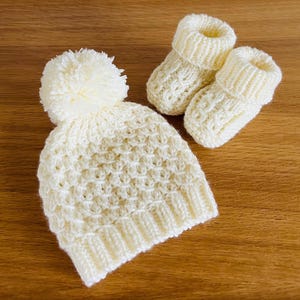 May include: A cream-colored, hand-knitted baby hat with a pom-pom and matching booties. The hat and booties are made of a soft, textured yarn. The set is displayed on a wooden surface.
