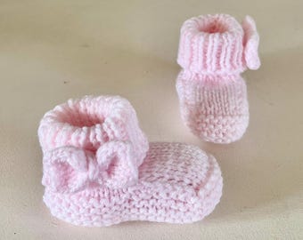 May include: A pair of soft pink knitted baby booties. One bootie features a bow detail, while the other has a folded cuff. The booties are made of a soft, textured yarn, perfect for a newborn.