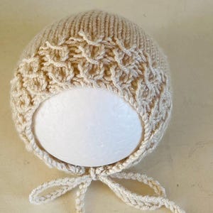 May include: A light beige knitted baby bonnet with a textured pattern on the crown. The bonnet has a rounded shape and a tie closure under the chin. The soft, neutral color makes it suitable for various outfits.