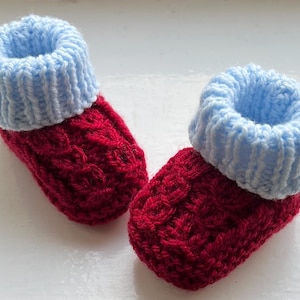 Può includere: Un paio di scarpine lavorate a maglia a mano con un motivo a trecce rosso scuro sul corpo principale e un polsino a coste azzurro a contrasto sulla parte superiore. Le scarpine sono realizzate in filato e presentano un design intrecciato e testurizzato.
