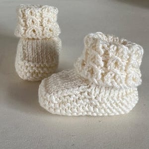 Könnte beinhalten: Ein Paar cremefarbene, handgehäkelte Babyschuhe. Die Schuhe haben einen umgeschlagenen Bund und ein strukturiertes Stichmuster. Das Garn scheint eine weiche, natürliche Faser zu sein, ideal für ein Neugeborenes.