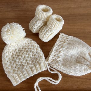 May include: A cream-colored, hand-knitted baby set featuring a hat with a pom-pom, a bonnet, and a pair of booties. The set is made of a textured knit pattern. The items are arranged on a wooden surface.