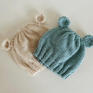 May include: Two knitted beanies, one in a light beige color and one in a muted blue color. Each beanie features a ribbed knit texture and two small, rounded ears attached to the top sides, creating a simple and functional headwear design.