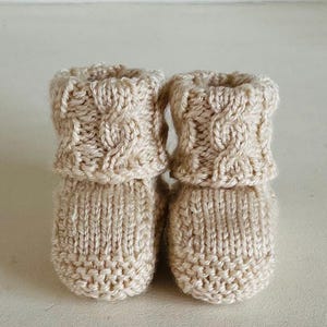 May include: A pair of beige knitted baby booties. The booties have a cable knit cuff and a ribbed knit foot. The yarn is a light, neutral color, and the booties appear soft and warm.
