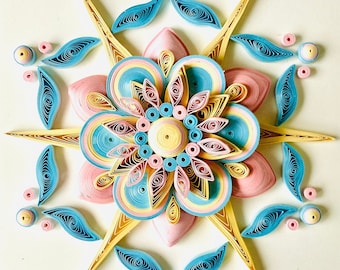 May include: A paper quilling art piece featuring a pink, yellow, and blue flower with intricate details. The flower is surrounded by smaller blue and yellow paper quilling designs.