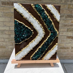 Op de afbeelding: Abstract kunstwerk op een klein ezel. Het kunstwerk heeft een donkerbruine achtergrond met diagonale banden van wit en smaragdgroen, omlijnd met goud. Het stuk is gemaakt van gemalen glas.