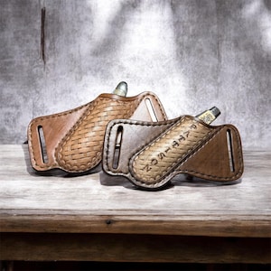 May include: Two brown and tan leather knife sheaths, featuring a woven pattern on the front. One sheath is embossed with the name "PATTISON". The sheaths, designed for knives, are displayed on a wooden surface.