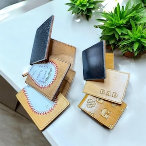 Puede incluir: Una colección de carteras de cuero en varios colores y diseños. Algunas carteras presentan costuras de béisbol, la palabra "DAD" y otros elementos decorativos. Las carteras están dispuestas sobre una superficie blanca, con plantas verdes en el fondo.