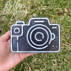 May include: Black and silver camera ornament with a textured surface. The camera has a circle in the center with a copyright symbol inside.
