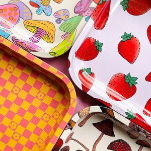 Puede incluir: Cuatro bandejas de metal diferentes con varios dise&ntilde;os. Una bandeja presenta un patr&oacute;n a cuadros en amarillo y rosa. Otra bandeja tiene un fondo blanco con un patr&oacute;n repetitivo de fresas rojas. Las dos bandejas restantes tienen un fondo blanco con un patr&oacute;n repetitivo de setas coloridas.