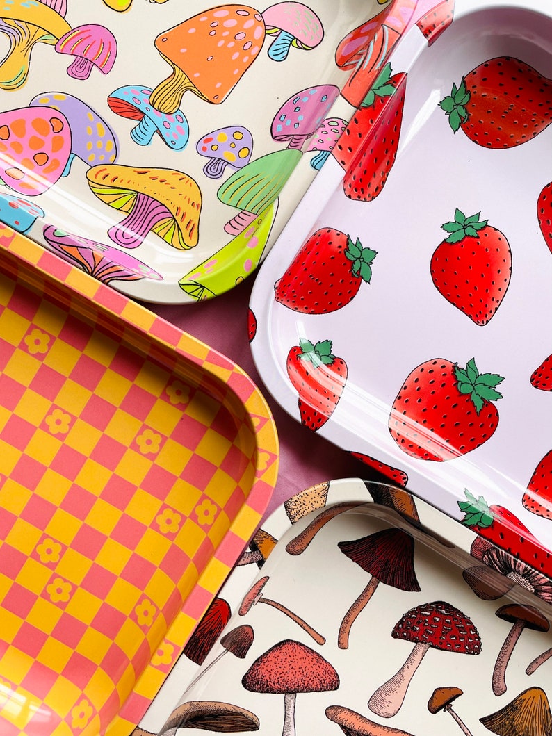 Puede incluir: Cuatro bandejas de metal diferentes con varios dise&ntilde;os. Una bandeja presenta un patr&oacute;n a cuadros en amarillo y rosa. Otra bandeja tiene un fondo blanco con un patr&oacute;n repetitivo de fresas rojas. Las dos bandejas restantes tienen un fondo blanco con un patr&oacute;n repetitivo de setas coloridas.