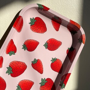 Puede incluir: Una bandeja de metal rosa con un patr&oacute;n repetido de fresas rojas. Las fresas tienen hojas verdes y est&aacute;n delineadas en negro.