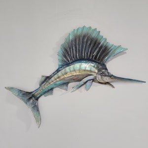 May include: A blue and green metal marlin fish wall sculpture with a long, pointed bill and a large dorsal fin. The fish is mounted on a white wall.