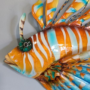 May include: A colorful metal sculpture of a lionfish with orange, white, and blue stripes. The fish has a large, fan-like tail and a prominent dorsal fin.
