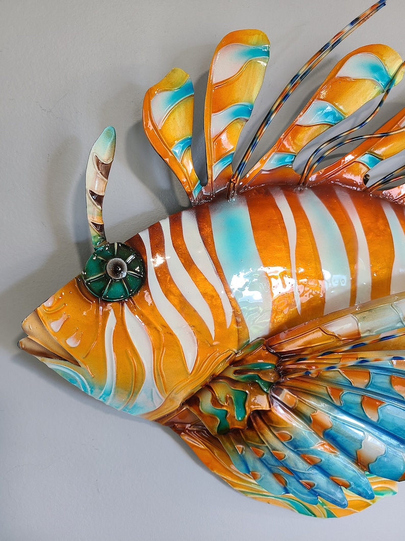 May include: A colorful metal sculpture of a lionfish with orange, white, and blue stripes. The fish has a large, fan-like tail and a prominent dorsal fin.