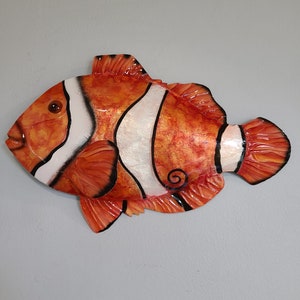 May include: A decorative clownfish wall hanging. The fish is orange with white stripes outlined in black. The fins and tail are detailed with a textured appearance. The fish has a dark eye and a curled design on its side.