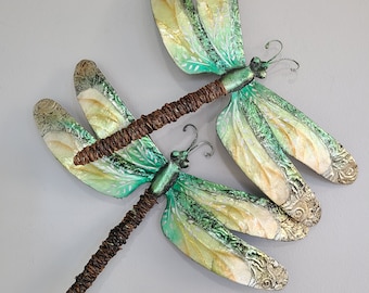 May include: Two green and yellow metal dragonflies with brown wire bodies. The dragonflies are attached to each other and are mounted on a white wall.