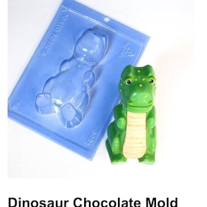 May include: A blue plastic dinosaur chocolate mold with a green plastic dinosaur next to it. The mold is shaped like a dinosaur sitting down with its legs crossed. The green dinosaur is also sitting down with its legs crossed.