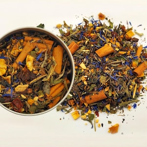 Puede incluir: Una lata de metal llena de té de hojas sueltas, junto a un montón disperso del mismo té. La mezcla de té incluye palitos de canela, trozos de fruta seca, acianos azules y hojas de té verde. El té es una mezcla de marrón, verde, amarillo y azul.