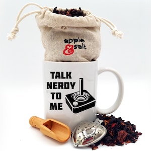 May include: A white ceramic mug with the text "TALK NERDY TO ME" and a joystick graphic. A small burlap bag with "apple & salt" text sits atop the mug. A wooden scoop and a heart-shaped tea infuser are next to the mug, with loose tea leaves.