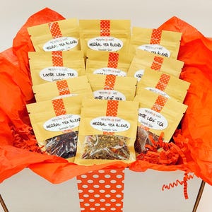 May include: A gift box filled with small tea sample bags. The bags are tan with a red and white polka dot ribbon. The box is lined with orange tissue paper and red shredded paper. The bags are labeled "Herbal Tea Blend" and "Loose Leaf Tea".