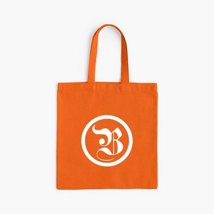 Op de afbeelding: Oranje draagtas met een wit cirkelvormig logo met een gestileerde letter "B". De tas heeft een rechthoekige vorm met een handvat om te dragen. De achtergrond is effen wit.