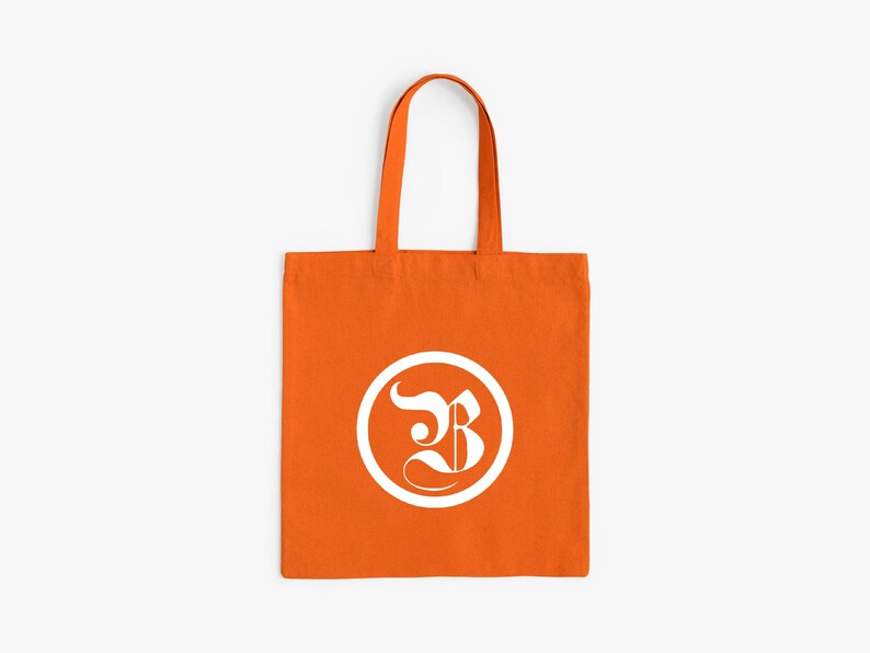 Op de afbeelding: Oranje draagtas met een wit cirkelvormig logo met een gestileerde letter "B". De tas heeft een rechthoekige vorm met een handvat om te dragen. De achtergrond is effen wit.