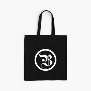 Op de afbeelding: Zwarte draagtas met een wit cirkelvormig logo met een gestileerde letter "B". De tas heeft een rechthoekige vorm met een handvat om te dragen. Het ontwerp is eenvoudig en modern, geschikt voor dagelijks gebruik.