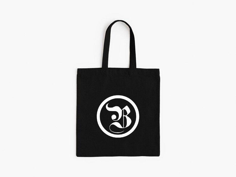 Op de afbeelding: Zwarte draagtas met een wit cirkelvormig logo met een gestileerde letter "B". De tas heeft een rechthoekige vorm met een handvat om te dragen. Het ontwerp is eenvoudig en modern, geschikt voor dagelijks gebruik.