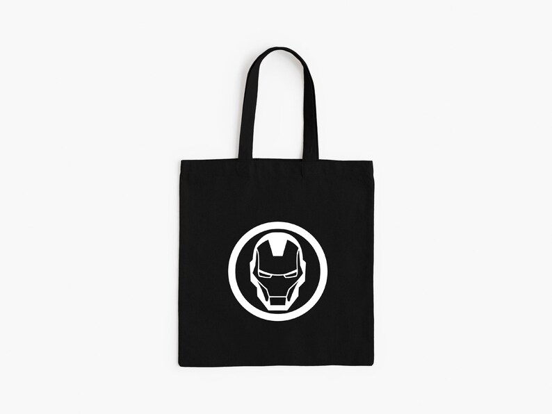 May include: Black tote bag featuring a white Iron Man helmet logo inside a white circle. The bag has black handles and is made of a durable fabric. The design is simple and bold, perfect for carrying books or groceries.